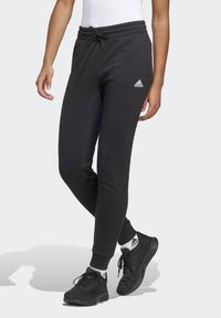 Fekete melegítőnadrág, húzózsinórral ellátott derékpánttal, szűkített lábszárakkal és egy kis fehér Adidas logóval a bal combon. Puha anyagból készült, bordás mandzsettákkal.