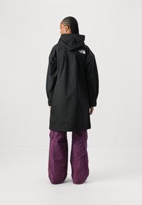 Veste imperméable noire avec capuche et logo dans le dos, associée à un pantalon violet ample. Présente une coupe large et des manches longues.