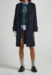 Persona che indossa un cappotto scuro sopra una felpa verde, una gonna di jeans e scarpe nere chunky, con guanti beige e calze fino al ginocchio.
