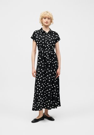 Γυναίκα με κοντά, σγουρά ξανθά μαλλιά, που φοράει ένα μαύρο, κοντομάνικο, μακρύ φόρεμα με κουμπιά και πουά σχέδιο, ζώνη στη μέση και μαύρες μπαλαρίνες.