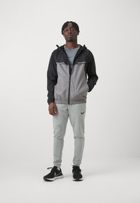 Veste zippée noire et grise avec taille élastique, associée à un pantalon de jogging gris clair avec un logo, et des chaussures de sport noires.