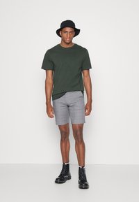 Donkergroen katoenen t-shirt, grijs geruite shorts, zwarte bucket hat en zwarte veterlaarzen. Eenvoudig ontwerp met een relaxte pasvorm.