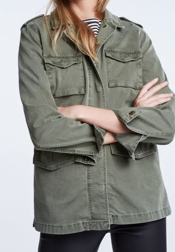 Personne portant une veste utilitaire vert olive avec de grandes poches sur le devant, sur une chemise rayée, bras croisés, seule la partie supérieure du corps est visible.