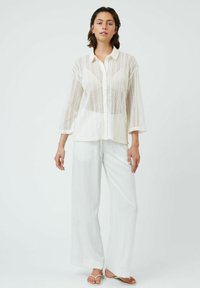 White, lightweight button-up shirt with a mesh pattern and three-quarter sleeves. Paired with loose, white linen pants and sandals.