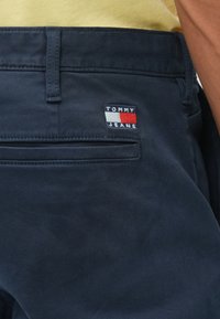 Marineblå bukser med flad front, baglomme med et rødt, hvidt og blåt "TOMMY JEANS" logo mærke, samt et slidstærkt tekstil.