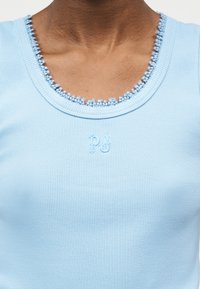 Κοντινό πλάνο ενός ατόμου που φοράει ανοιχτό μπλε πλεκτό τοπ με κυματιστό ντεκολτέ και μικρά κεντημένα γράμματα "p" και "j" στο στήθος.