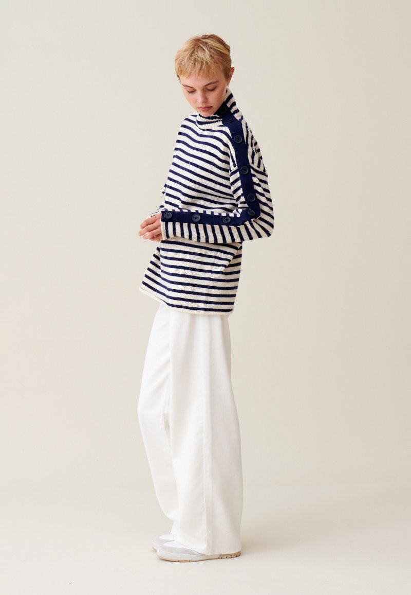 Gestreifter Pullover in Marineblau und Weiß mit Rollkragen, Knopfakzenten entlang eines Ärmels, kombiniert mit weiten weißen Hosen.