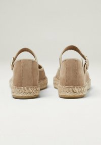 Chaussures espadrilles en suède beige avec une semelle en jute tissée, dotées d'une sangle à la cheville et d'une boucle métallique décorative sur le talon.