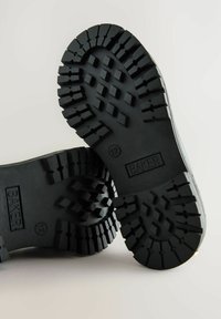 Schwarze Gummistiefel-Sohlen mit tiefen Profilmustern. Auf der Seite sind das Branding "BAKER" und die Größe "12" geprägt.