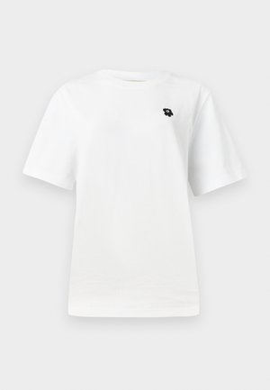 Hvit t-skjorte med korte ermer som har et lite svart grafisk motiv på venstre brystområde, satt mot en enkel lys bakgrunn.