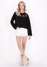 Maglia nera a maniche lunghe con ricami floreali sul collo, abbinata a pantaloncini in denim bianchi strappati e stivaletti beige alla caviglia.