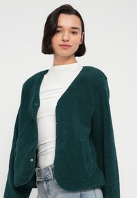 Τζάκετ από φλις σε θαλασσί χρώμα με απαλή, υφή, που χαρακτηρίζεται από χαλαρή εφαρμογή, ανοιχτό μπροστινό μέρος και διακριτικές λεπτομέρειες με κουμπιά. Φοριέται πάνω από λευκό τοπ.