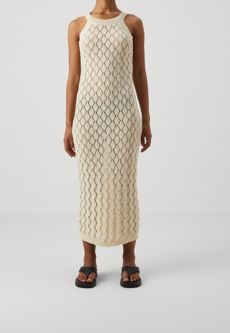Femme portant une robe midi sans manches, en maille ouverte crème avec un motif en losange et des sandales noires sur un fond uni.