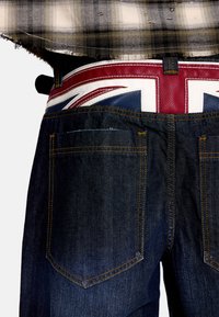 Σκούρο τζιν με κίτρινα ραφές, με δερμάτινη ζώνη στη μέση σε χρώματα κόκκινο, άσπρο και μπλε με το Union Jack. Δύο πίσω τσέπες είναι ορατές.