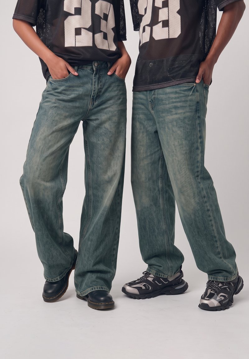 Deux personnes portant des maillots de sport noirs assortis et des jeans bleus délavés et amples, debout côte à côte les mains dans les poches.