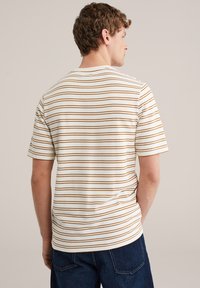 Ung mand set bagfra iført en hvid t-shirt med korte ærmer og tynde brune vandrette striber samt mørkeblå jeans mod en ensfarvet baggrund.