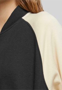 Svart og kremfarget sweatshirt med ribbestrikket hals, mykt stoff og et delt fargedesign med kontrasterende teksturer på ermene.