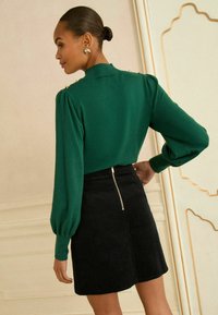 Zaļa gara piedurkņu blūze ar pūkām uz pleciem, ar zelta akcentiem plecu daļā. Saskaņota ar melnu kārni jostu, kas aprīkota ar aizmugurējo rāvējslēdzēju.