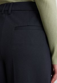 Dunkelblaue Hosen mit glatter Textur, ausgestattet mit einem Detail am Gesäßtasche. Getragen mit einem leicht olivfarbenen, gerippten Langarmoberteil.