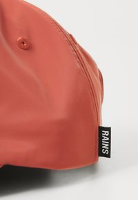 Gros plan sur un capuchon orange texturé avec une étiquette noire portant l'inscription "RAINS" près du bord, mettant en avant son design moderne.