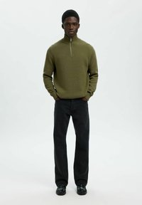 Alie žalios spalvos, ilgomis rankovėmis, užtrauktuku sudedamas megztinis su raukšlėtu paviršiumi. Su juodais džinsais ir juodais batais, stovintis prieš šviesų foną.