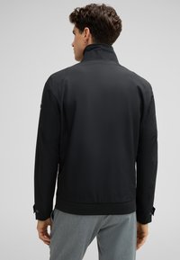 Schwarze Jacke mit hohem Kragen und geripptem Saum, mit glatter Textur und geknöpften Manschetten, von hinten betrachtet.