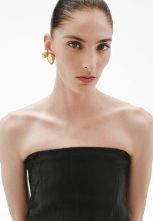 Top negro sin tirantes con tejido texturizado. La modelo lleva pendientes llamativos de oro. Piel lisa y cabello liso, estilo minimalista.