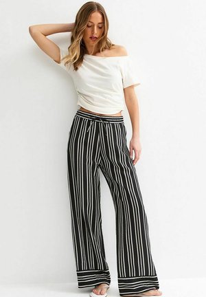Mujer con blusa blanca de hombros descubiertos y pantalones anchos negros con rayas verticales blancas, de pie junto a una pared blanca.