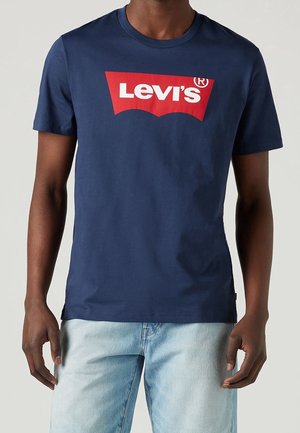 Homme portant un t-shirt Levi's bleu marine avec un logo rouge et blanc et un jean bleu clair, debout devant un fond uni.