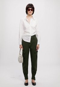 Weißes Hemd mit Knöpfen, olivgrüne Cargo-Hosen mit Taschen, schwarze Ballerinas und eine kleine hellgraue Handtasche. Schwarze Sonnenbrille runden das Outfit ab.