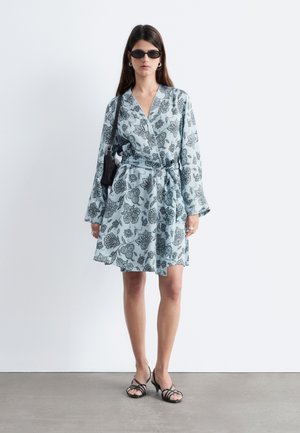 Žena u svijetloplavoj cvjetnoj haljini na preklop, crnim sandalama s remenčićima, crnoj torbici preko ramena i tamnim sunčanim naočalama na čistoj bijeloj pozadini.