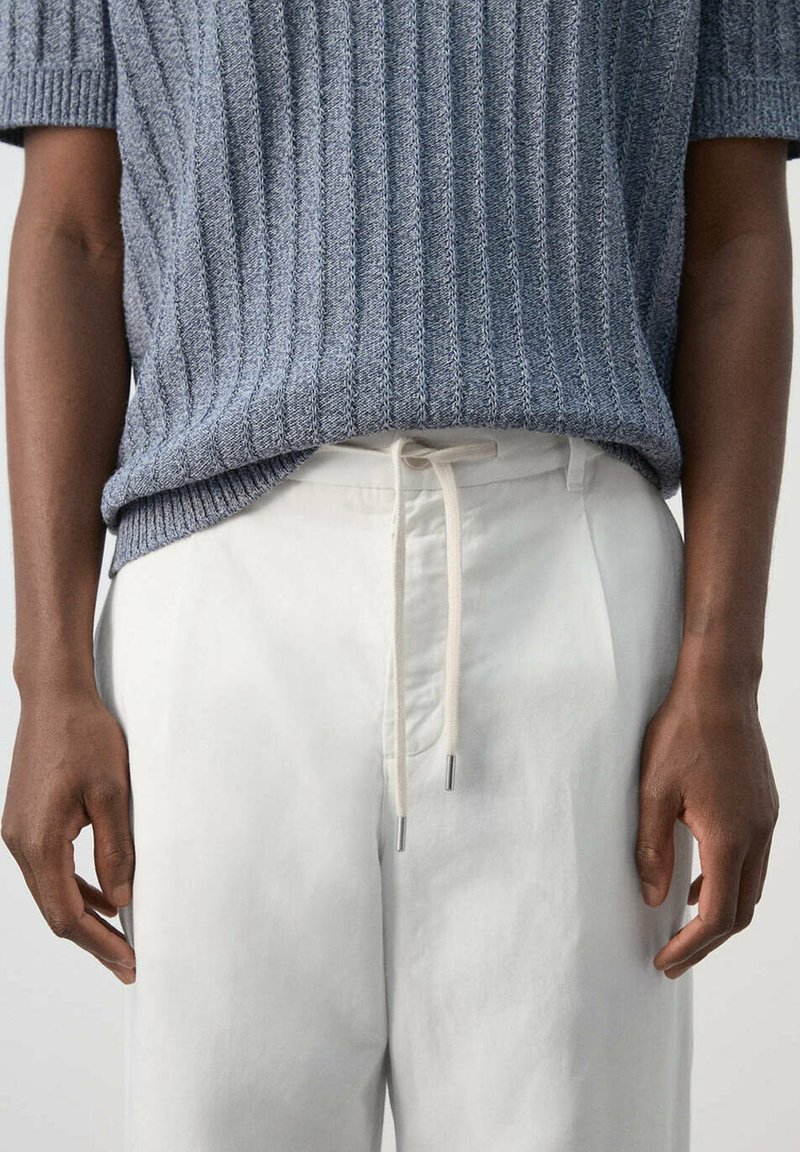 Gros plan d'une personne portant un pull en tricot gris clair à manches courtes, rentré dans un pantalon blanc à cordon, les mains détendues sur les côtés.