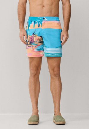 Hombre que lleva pantalones cortos de baño coloridos con estampado de playa y edificios, combinados con alpargatas verdes sin cordones, de pie frente a un fondo liso.