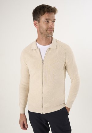 Beige geribde gebreide cardigan met een ritssluiting aan de voorkant, een kraagontwerp en aansluitende mouwen. Draagt over een wit shirt en gecombineerd met donkere broek.