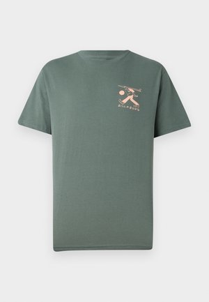 Maglietta verde a maniche corte con una piccola grafica di una pesca di una persona che trasporta una tavola da surf e la parola "Billabong" sul petto sinistro.