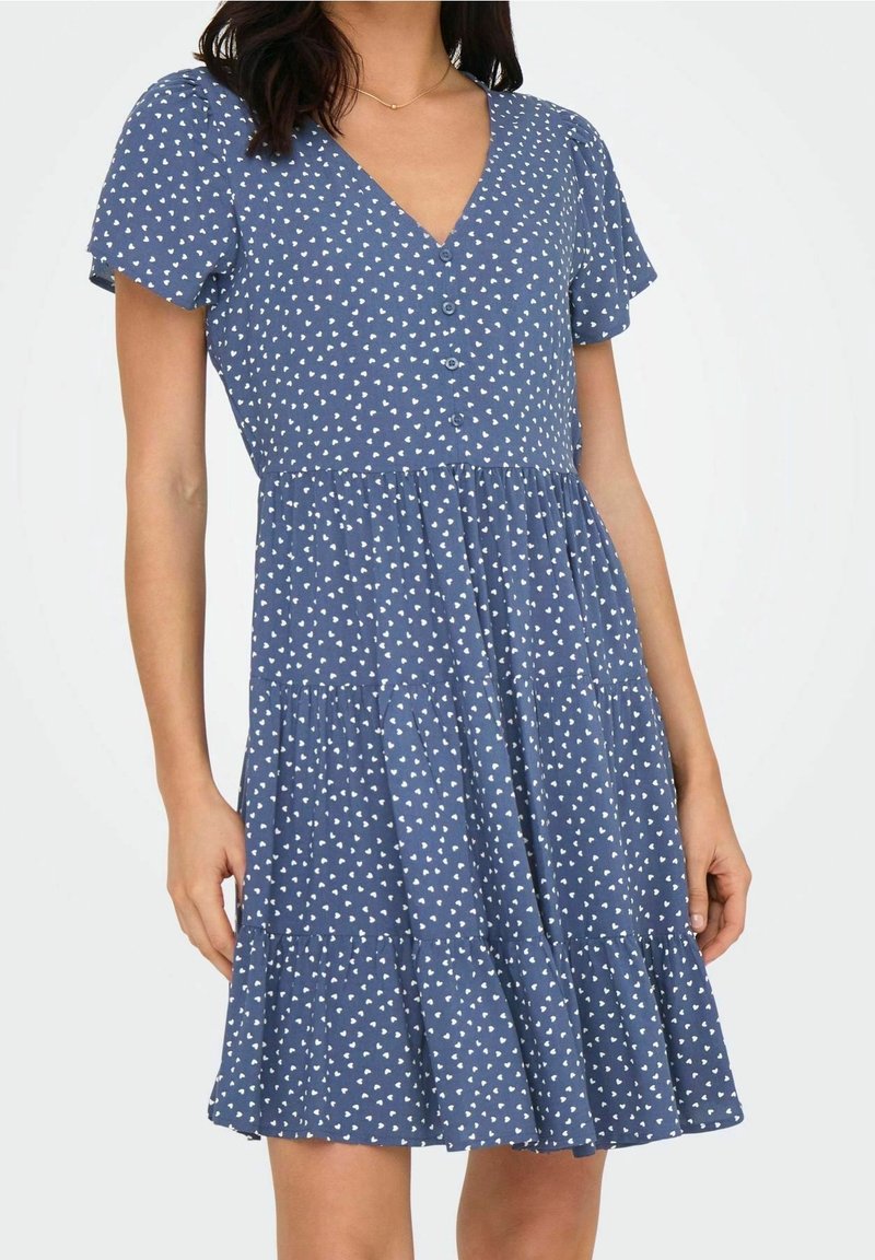 Femme portant une robe courte à manches courtes bleue avec des motifs de cœurs blancs, des boutons sur le devant et une jupe à volants en plusieurs niveaux.