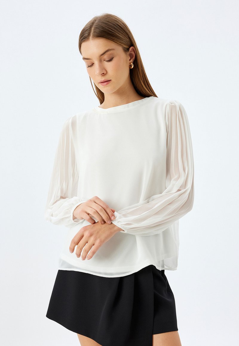 Blusa bianca con maniche lunghe e trasparenti a palloncino. Tessuto morbido, scollatura rotonda e polsini arricciati. Abbinata a una gonna nera.