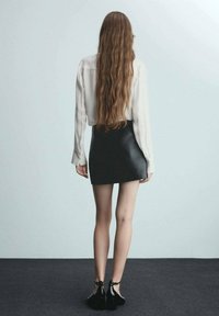 Černá kožená mini sukně v kombinaci s bílou blůzou s dlouhým rukávem. Modelka má dlouhé, vlnité vlasy a nosí černé platformové boty s kotníkovými šněrováním.