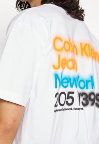 Biała koszulka z nadrukiem graficznym z napisem "Calvin Klein Jeans", "New York" oraz szczegółami liczbowymi w kolorach pomarańczowym, niebieskim i czarnym. Miękki bawełniany materiał.