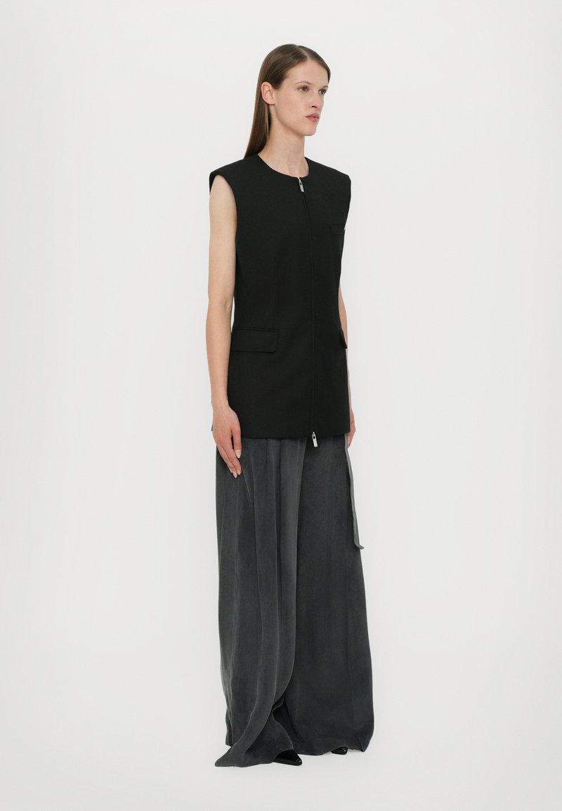 Black sleeveless vest with a front zipper, two pockets, and a structured design, paired with loose, gray pants in a soft, draping fabric.