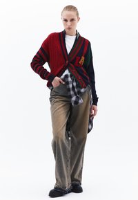 Stickad kofta i röd, grön och marin med kabelmönster; i kombination med rutig skjorta, löst sittande grå jeans och svarta skor.