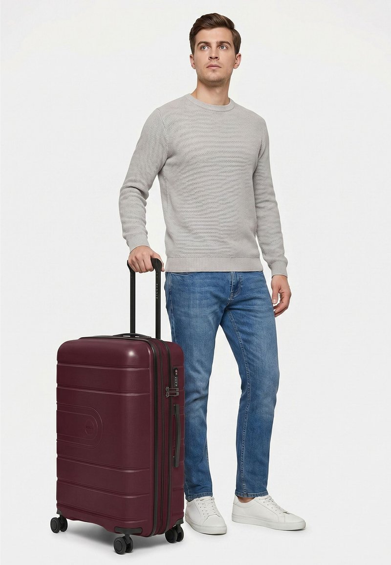 Giovane uomo con maglione grigio e jeans blu in piedi che tiene la maniglia di una grande valigia con ruote color amaranto.
