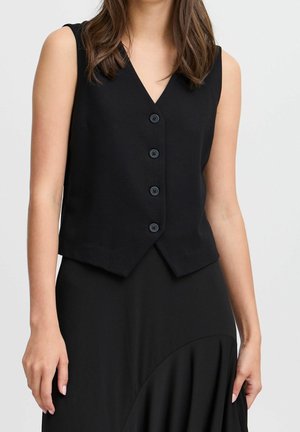 Femme portant un gilet noir sans manches à boutons et une jupe noire avec un détail froncé, debout devant un fond clair uni.