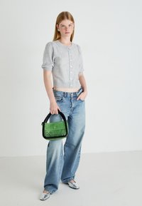Jonge vrouw in een grijze kortmouwige gebottonde trui, wijde jeans, zilveren schoenen, met een groene en zwarte kraaltas.