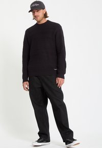 Sort strik sweater med struktureret mønster og rund hals, parret med løstsiddende mørke jeans og sorte lavtoppede sneakers. Hat med logo.