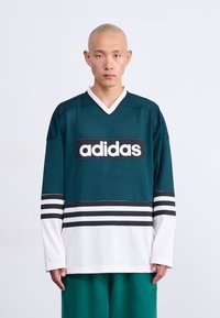 Muškarac s obrijanom glavom nosi zelenu i bijelu Adidas sportsku majicu i zelene hlače, stoji naslonjen na jednostavnu bijelu pozadinu.