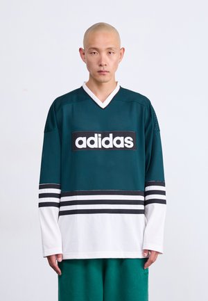 Mann mit rasiertem Kopf, der ein grün-weißes Adidas-Sporttrikot und grüne Hosen trägt, steht vor einem schlichten weißen Hintergrund.