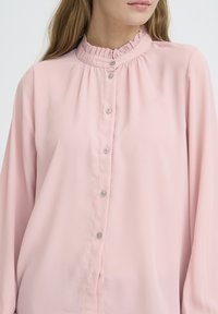Light pink blouse with a ruffled collar, long sleeves, and button closure. Smooth fabric with a soft texture and subtle sheen.