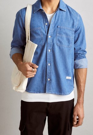 Man draagt een blauwe spijkerblouse over een wit T-shirt, zwarte broek, en draagt een beige canvas tas over de schouder.