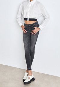 Persona que lleva una camisa blanca corta con botones, unos jeans ajustados negros de cintura alta y zapatos de plataforma blancos gruesos con recortes.